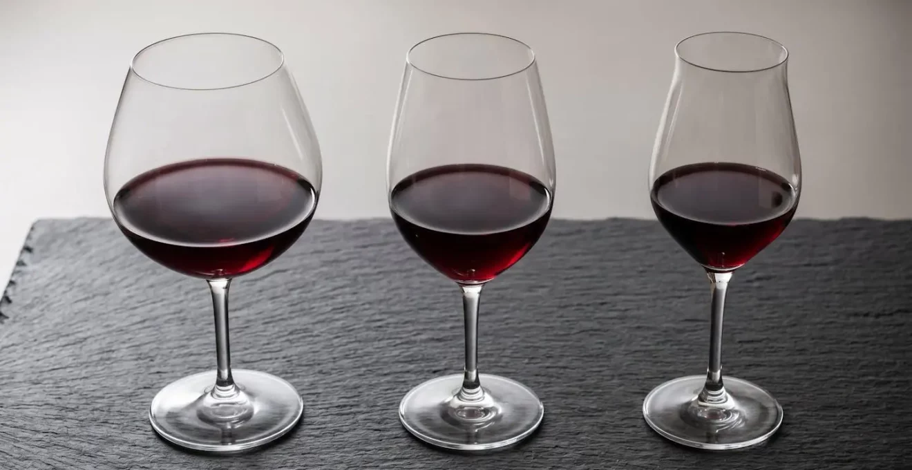 Trois verres à vin de formes différentes (ballon, tulipe, intermédiaire) alignés côte à côte, remplis du même vin rouge, montrant les différences de surface de contact et d'ouverture